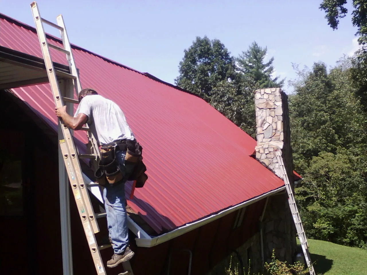 Roofer on a ladder beside a red metal roof and stone chimney — Soffit & Fascia Repair by Roofing Brothers in Coalville